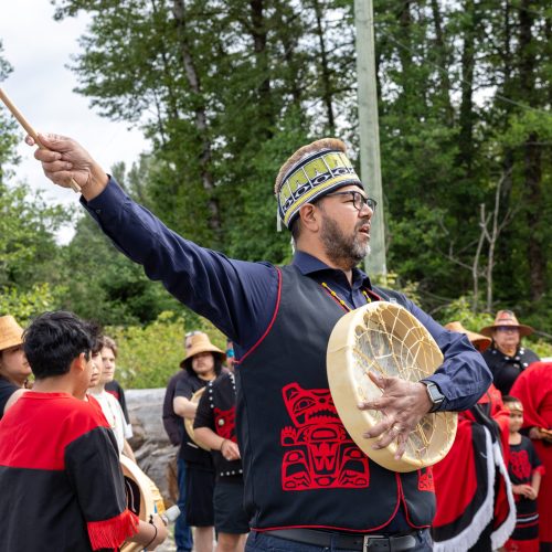 A man in traditional wear plays a drum outdoors, surrounded by a group in similar clothing.